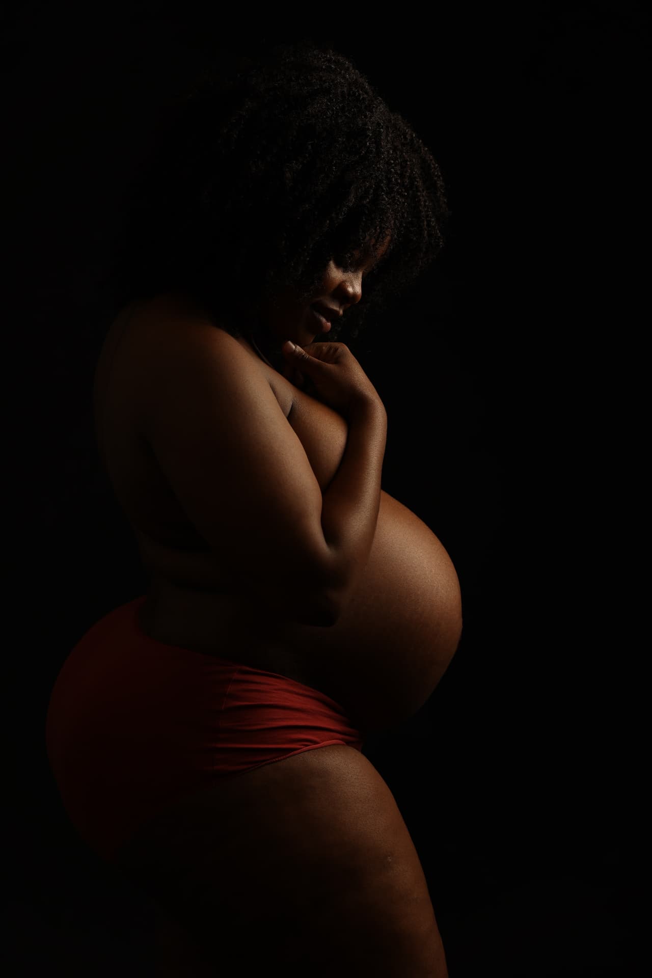 Dramatic low-key studio maternity portrait in profile, the mother-to-be cradling her bump against a black background