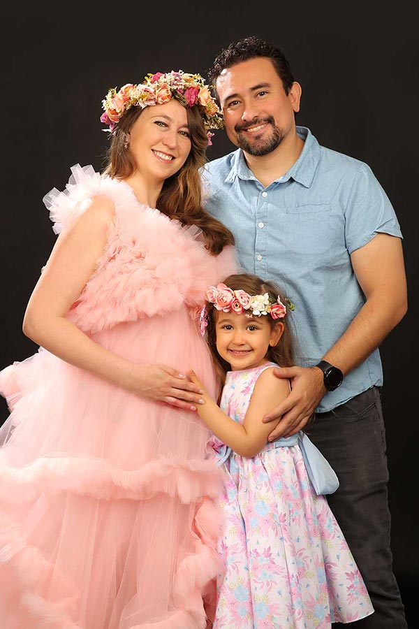 Expectant mother in a pink tulle gown with floral crown, her partner and daughter resting their hands on her baby bump in the studio