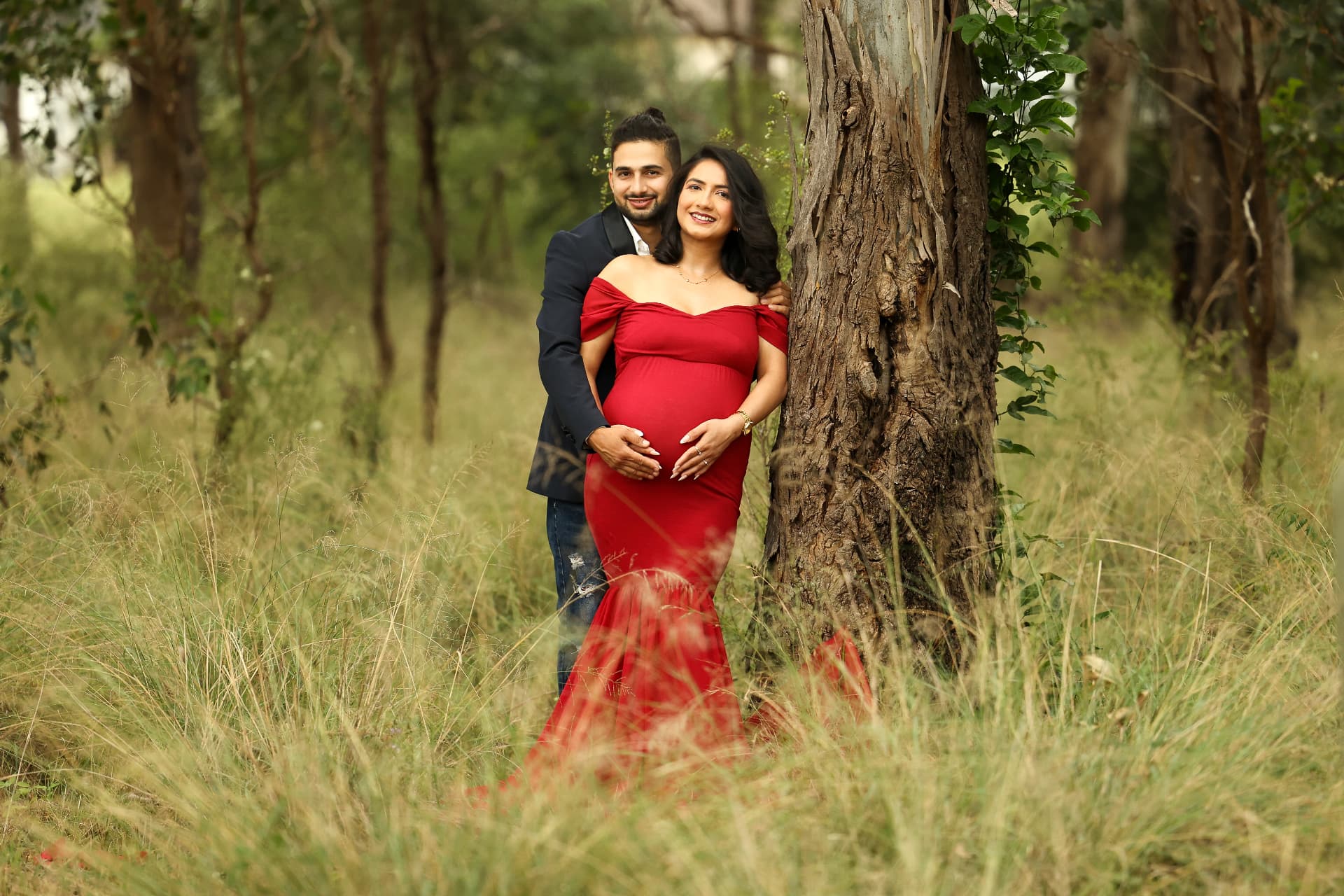 Expectant couple embracing outdoors in a red mermaid gown among tall grass and trees