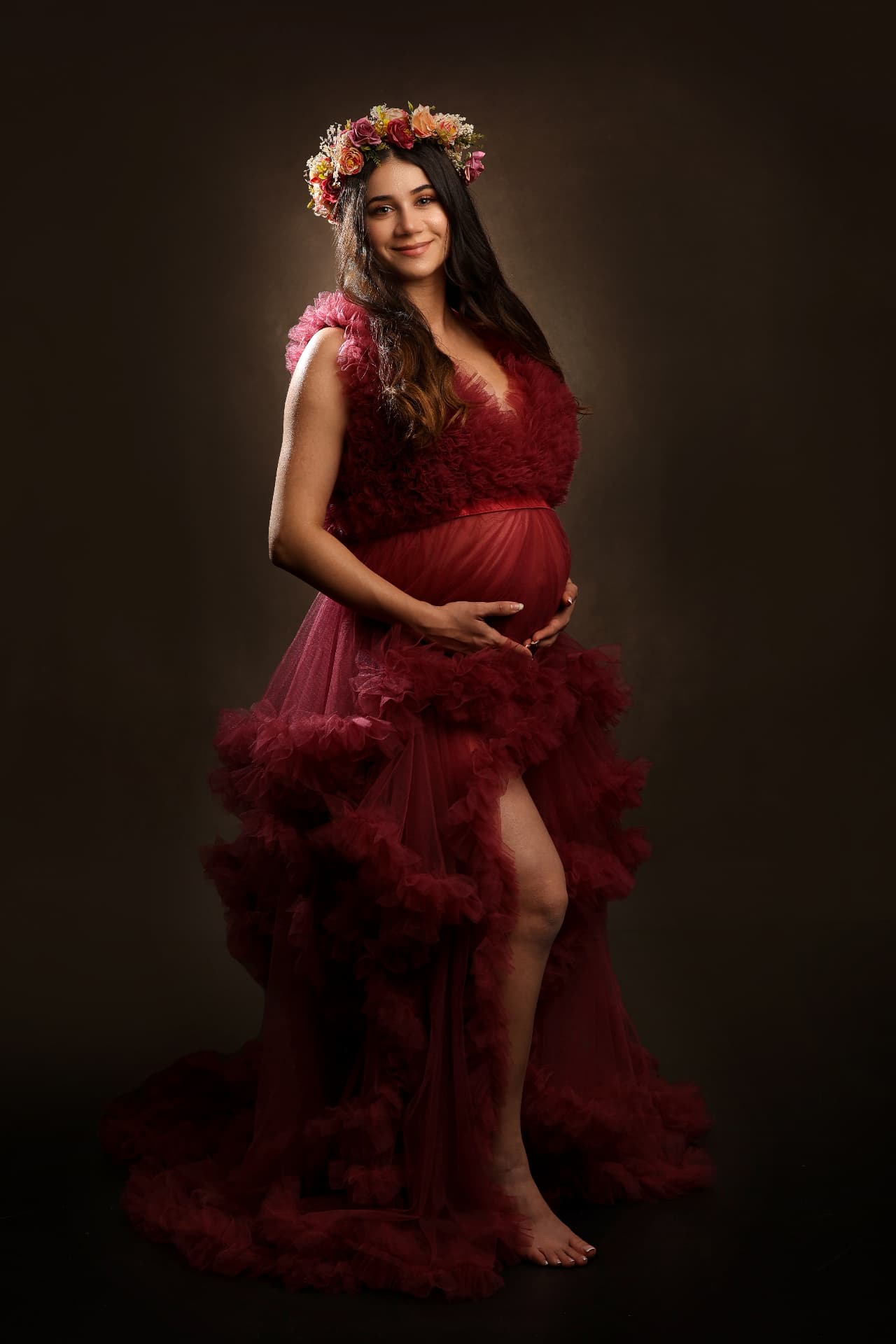 Expectant mother in a deep burgundy ruffled tulle gown with a floral crown, cradling her bump under dramatic studio spotlighting